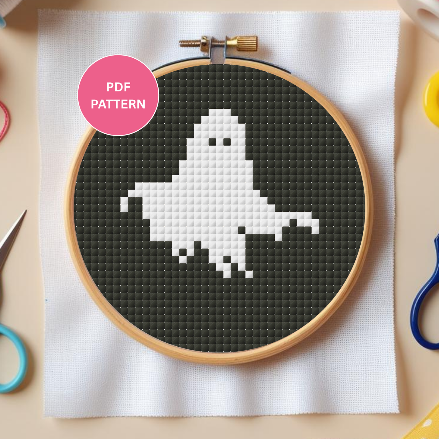 Cross-stitch hoop with a ghost design on dark fabric, surrounded by crafting tools.
